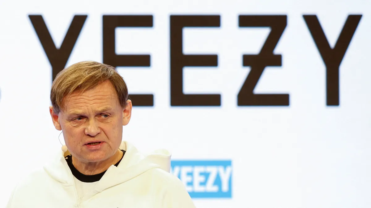 Adidas CEO Bjørn Gulden speaking in front of a white background with a Yeezy logo on it.