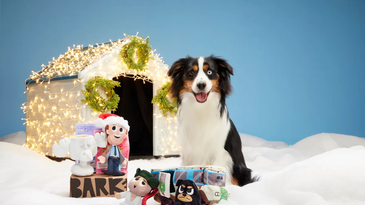 A dog sitting in front of a dog house with Christmas lights on it and dog toys in front of it.