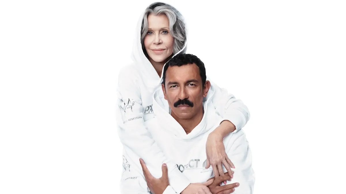 Two people, both wearing white, one in a hoodie, the other a mustache, embrace.