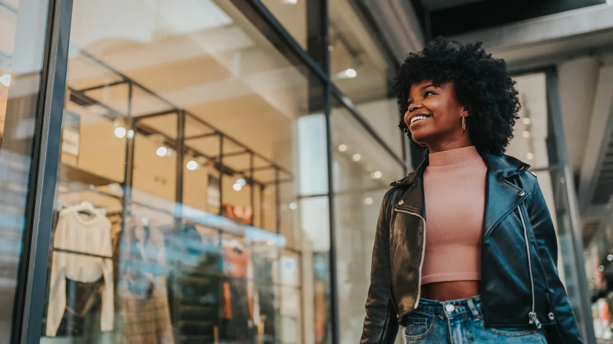 A smiling person in a black leather jacket walks by a clothing store window.