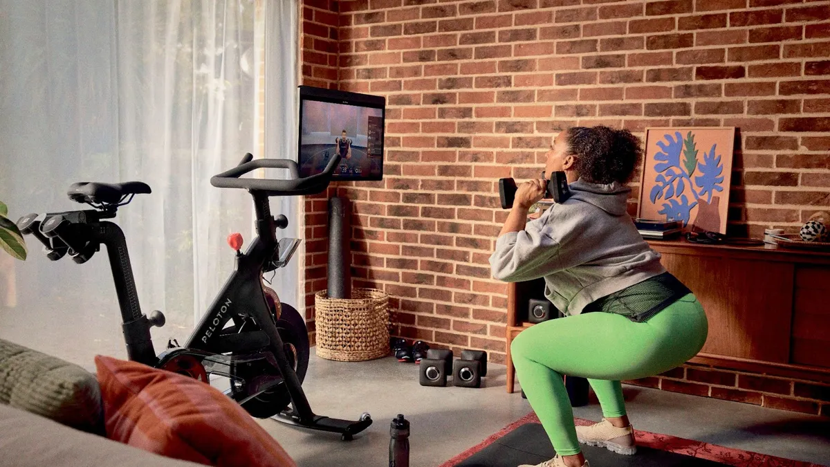 A person doing a squat while holding weights and following an instructional Peloton video, with the exercise brand's Peloton Bike also in view.