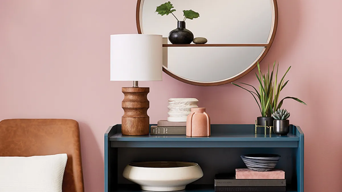 A round mirror with a small shelf holding a plant hangs above a blue bookcase. Items there include a lamp, another plant, books and decorative bowls. A bed with a white pillow is seen on the left.