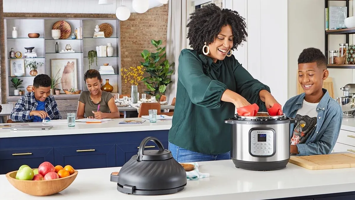 Two kids do homework while an adult and another child use a kitchen appliance.