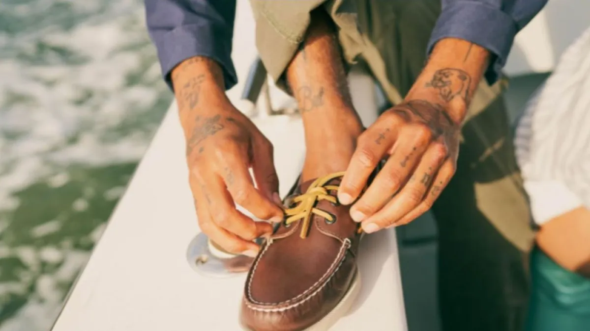 Person with hand tattoos kneels and laces up a Sperry boat shoe.